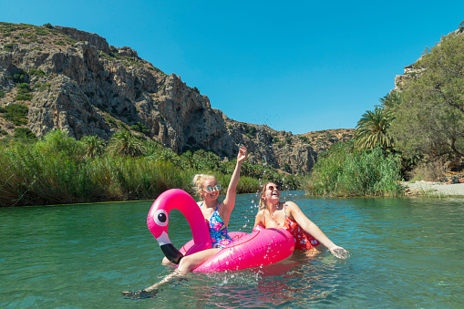 Women friends sitting in a flamingo pool float on a lake. One has he arm up in the air while they both laugh.