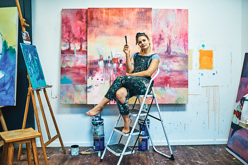 Full length portrait of an attractive young woman sitting on a ladder during a painting session in her art studio
