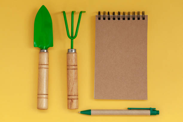 Wooden handled garden hand trowel and hand fork and empty noteboook on yellow background