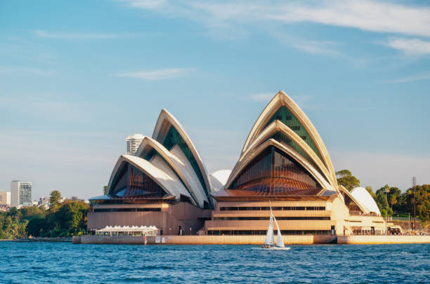 Sydney Australia - August 5, 2017: A sunny day in Sydney, Australia, and a small sailing boat crosses the blue waters of Port Jackson. Beyond it can be seen the iconic shape of the Opera House.