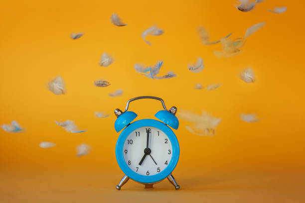 istock Soft fluffy flying feathers and blue alarm clock on yellow background. 1210531827
