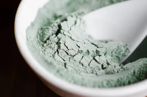 Photo of Green bentonite clay powder in a bowl. Diy facial mask and body wrap recipe. Natural beauty treatment and spa. Clay texture closeup, selective focus.