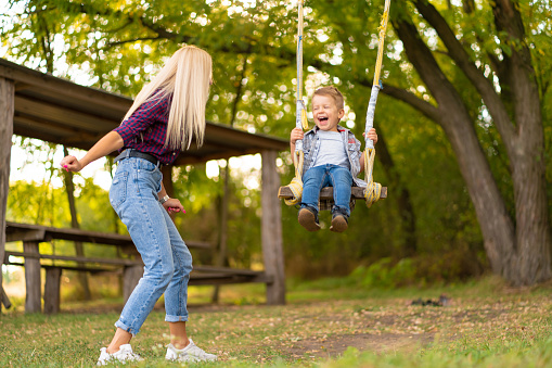 Young blonde mom shakes her little son on a swing in a green park. Happy childhood