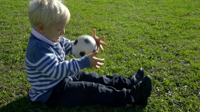 three year old boy sitting on the green grass on a sunny day with a soccer ball