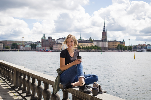 Modern young woman sitting on a fence in front of colorful Gamla Stan in Sweden