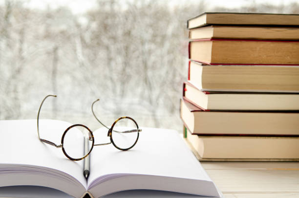 istock Round reading glasses lie on an open book with white pages next to a stack of multi-colored books on a windowsill 1210551263