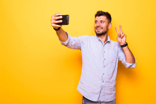 istock Portrait of a smiling bearded man showing peace gesture while taking a selfie isolated over yellow background 1210551940