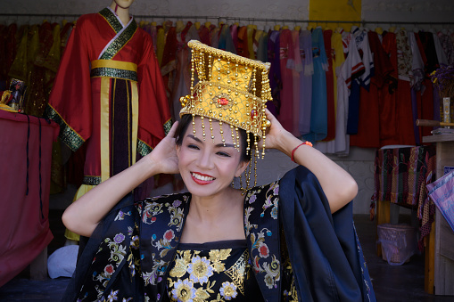 Woman wearing traditional Chinese dress with head gear smiling with all style and color hang in side room background