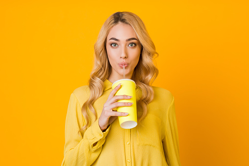 Young woman drinking juice from paper cup, standing over yellow studio background