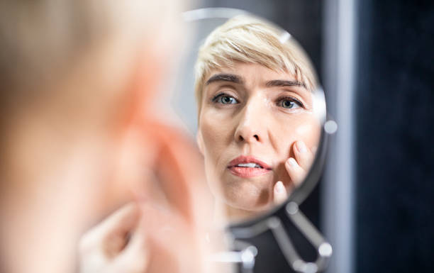 femme mûr regardant dans le visage touchant de miroir restant dans la salle de bains - aging process photos photos et images de collection