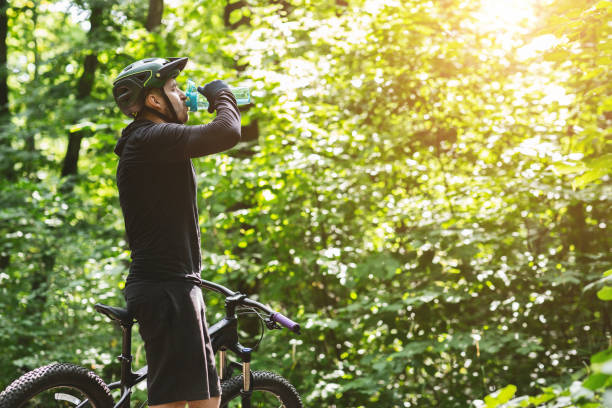 istock Young athletic cyclist drinking water over green forest background 1210557992
