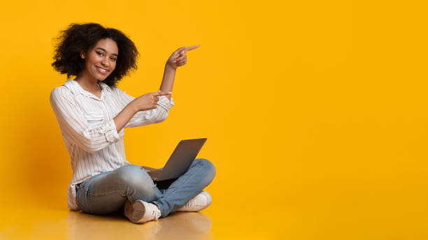 Cheerful Afro Girl Sitting On Floor With Laptop And Pointing Aside Online Offer. Cheerful Afro Girl Sitting On Floor With Laptop And Pointing Away With Fingers, Indicating Copy Space On Yellow Background, Panorama girl sitting stock pictures, royalty-free photos & images