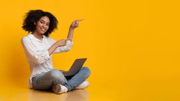 Photo of Cheerful Afro Girl Sitting On Floor With Laptop And Pointing Aside