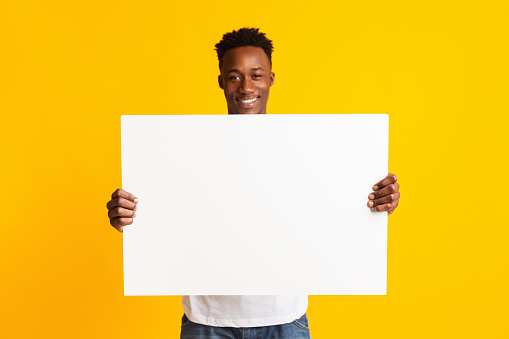 Look here. Excited african american man holding blank poster with copy space on yellow studio background