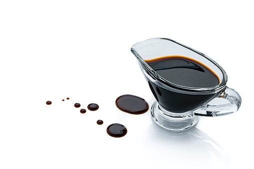 High angle view of balsamic vinegar in a gravy boat isolated on reflective white background. Some vinegar drops are spilled on the background beside the container. The composition is at the right of an horizontal frame leaving useful copy space for text and/or logo at the left. Predominant colors are black and white. High resolution 42Mp studio digital capture taken with Sony A7rII and Sony FE 90mm f2.8 macro G OSS lens