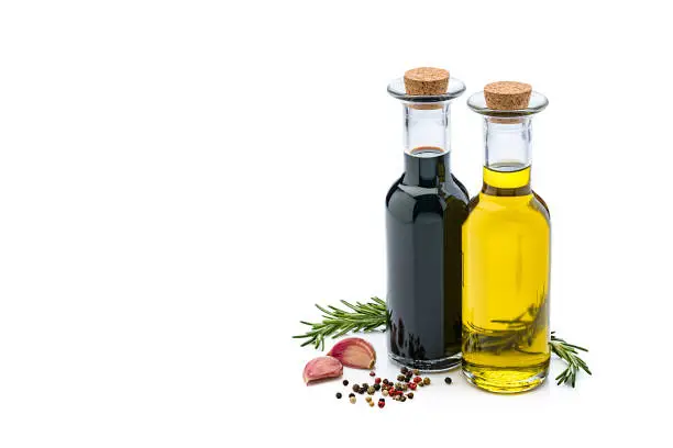 Extra virgin olive oil and balsamic vinegar bottles isolated on reflective white background. Rosemary twigs, garlic cloves, and mixed peppercorns are around the bottles and complete the composition. The composition is at the right of an horizontal frame leaving useful copy space for text and/or logo at the left. Predominant colors are yellow, black and white. High resolution 42Mp studio digital capture taken with Sony A7rII and Sony FE 90mm f2.8 macro G OSS lens