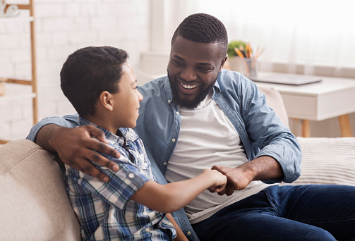 Father Son Bonding. Portrait Of Cheerful Black Boy Bumping Fists With His Dad, Sitting On Sofa At Home, Enjoying Spending Time Together