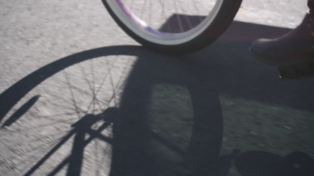 Slow Motion Shot of a Bicycle's Wheels and Spokes Casting a Shadow on Pavement while a Woman Wearing Leather Boots Pedals Outdoors on a Sunny Day