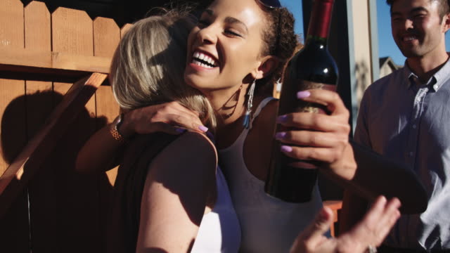 Slow Motion Shot of a Caucasian Woman in Her Forties with Blonde Hair Greeting a Young Woman of Mixed Race (Carrying a Bottle of Wine) and a Young Asian Man with Smiles and Hugs at the Gate of a Wooden Fence of a Home on a Sunny Day