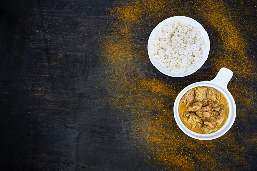 Orange spicy chicken curry and rice in simple white plates on a dark background. Around hot pepper, lemon, curry powder. Indian traditional cuisine. Flat lay. Top view, copy space