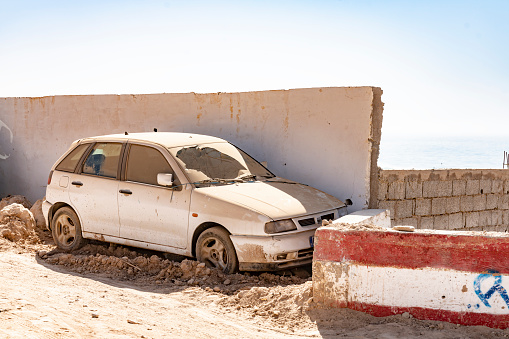 Old car with broken windscreen left at the side of the road. This is in Taghazout, Morocco and near a construction site. it is behind a wall but covered in sand as it has been there some time.