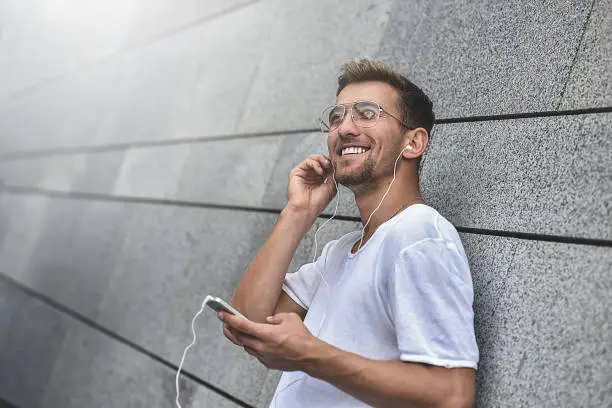 Photo of Cheerful guy dressed in white t-shirt at the street, listening to music with earphones, holding mobile phone.