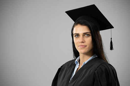 Portrait of a happy Caucasian female student wearing a gown and a hat graduating from high school college looking at camera on grey background.