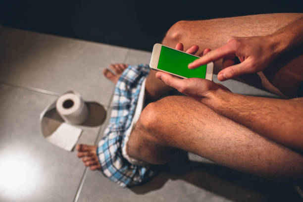 男はトイレやトイレのポットに座っています。男は、グリーンスクリーンで電話を保持し、それで遊びます。その上に指でポイントし、タップします。床にロール紙。足のブリーフ。排便。 - mobile phone smart phone toilet water ストックフォトと画像