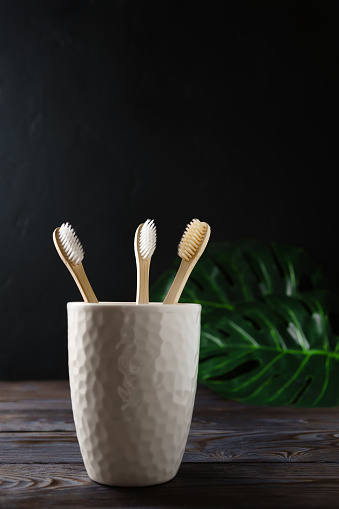 White ceramic glass with three biodegradable bamboo toothbrushes stands on a wooden table, dark background. Image with copy space. Zero waste concept.
