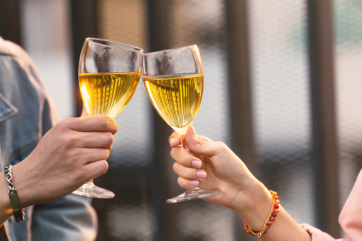 close up hands of lovers couple man and woman cheering and toast with white wine glasses to celebrating at a dinner party in the summertime. love, celebration, relationship and romantic concept.