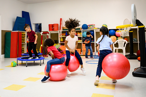 Active Hispanic schoolchildren bouncing on fitness balls and mini trampolines in school playroom.