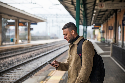 Handsome Tourist Watching Content on his Mobile Phone Waiting for Train to Come - Stock Photo