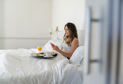 Happy woman at a hotel texting on her cell phone while having breakfast in bed - room service concepts