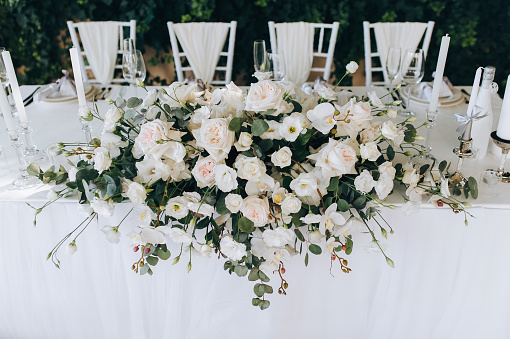 A wedding table with a white tablecloth, candles, a large bouquet of white roses and vintage chairs stands against a background of green foliage of wild grapes. Outdoor, summer ceremony. Close up.