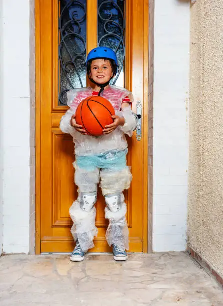 Boy with basketball ball wear super safe bubble wrap and helmet have overprotective mother standing near home door