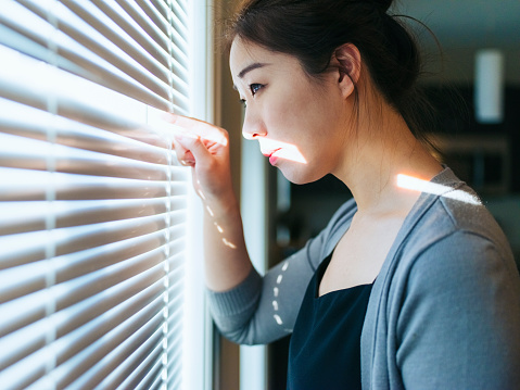 young woman looking out of blinds at home.