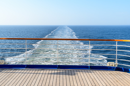 Aft deck of a wooden-coated cruise ship with railings. View of the wake waves behind the stern. Sea horizon in the ocean.