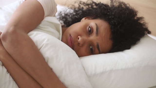 Depressed african woman lying on bed embraces pillow feels unhappy