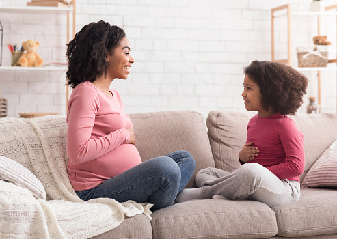 Expecting mom telling her little daughter about pregnancy, curious cute girl touching belly as mother, sitting on couch in living room, side view