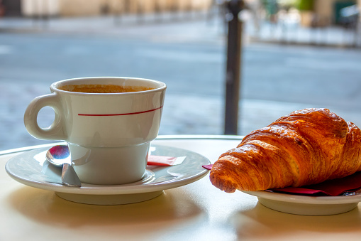 Little table of street cafe in Paris in the morning. Cup of coffee and croissant close-up. Background in defocus