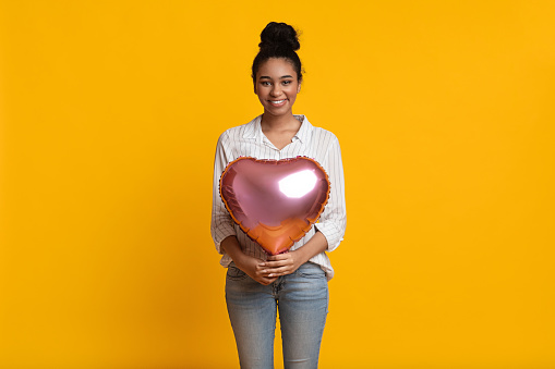 Symbol of love. Beautiful afro girl with heart shaped air balloon on yellow background. Happy black woman on Valentine's Day, Free Space