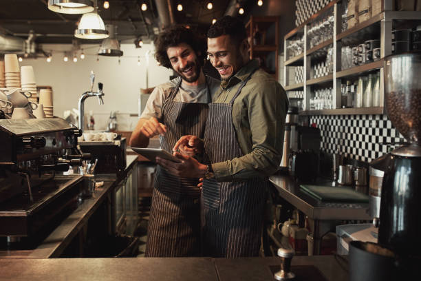 istock Smiling handsome waiter holding tablet with coworker while pointing on it in coffee shop sharing recipe 1210733599