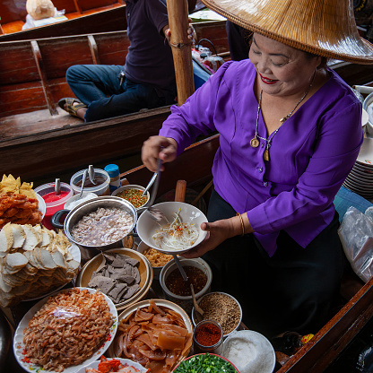 Traditional Thai noodle vendor, wearing a traditional straw hat, selling her freshly prepared traditional Thai noodle soup from a canal boat at Damnoen Saduak floating market. Many different fresh ingredients can be seen displayed around a large pot of boiling water, which is used to soften the fresh rice noodles and boil different meats.