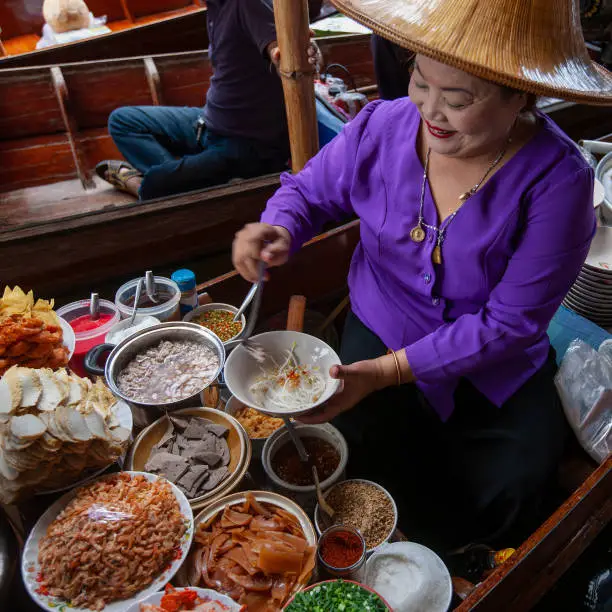Photo of Traditionally dressed Thai noodle vendor selling her freshly prepared traditional Thai noodle soup from a canal boat at Damnoen Saduak floating market.