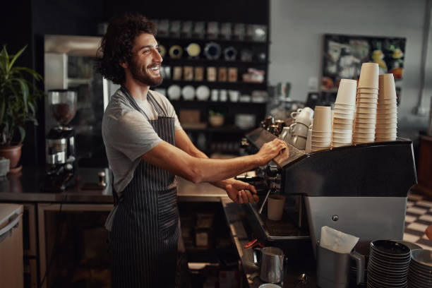istock Young cheerful barista wearing black apron while preparing coffee at an automatic machine in a modern coffee shop 1210733748