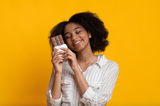 Portrait of happy afro girl hugging chocolate bar, standing with closed eyes over yellow background in studio, copy space
