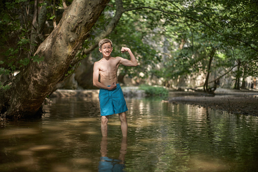 Smiling happy teenage boy having fun by posing like bodybuilder in shallow water in sunny forest in mediterranean country in summer time, relaxed mood fun smile happiness