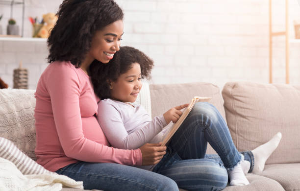 istock Pregnant afro mom and daughter spending time at home, reading book 1210744433