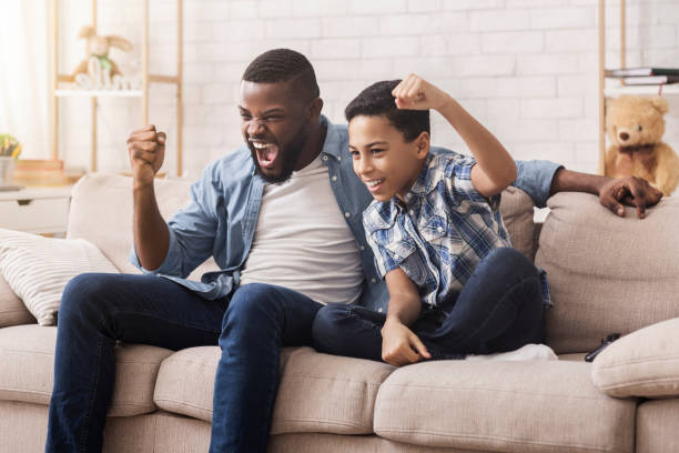 afro papà e figlio guardano lo sport in tv e tifano emotivamente - little boys people indoors soccer foto e immagini stock