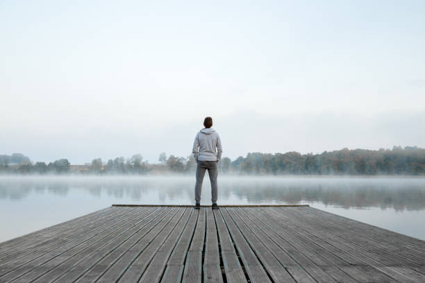 junger mann steht allein auf holzischem steg und starrt auf den see. denken über das leben. nebel über wasser. nebelige luft. früh kühler morgen. friedliche atmosphäre in der natur. genießen sie frische luft. rückansicht. - bootssteg stock-fotos und bilder